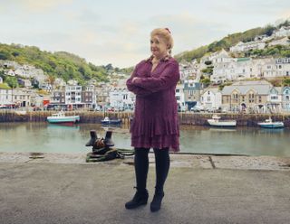 A shot of Felicity Montagu as Margo Martins in Beyond Paradise, standing in the harbour of Shipton Abbott with the ocean and the houses going up the cliffs in the background behind her