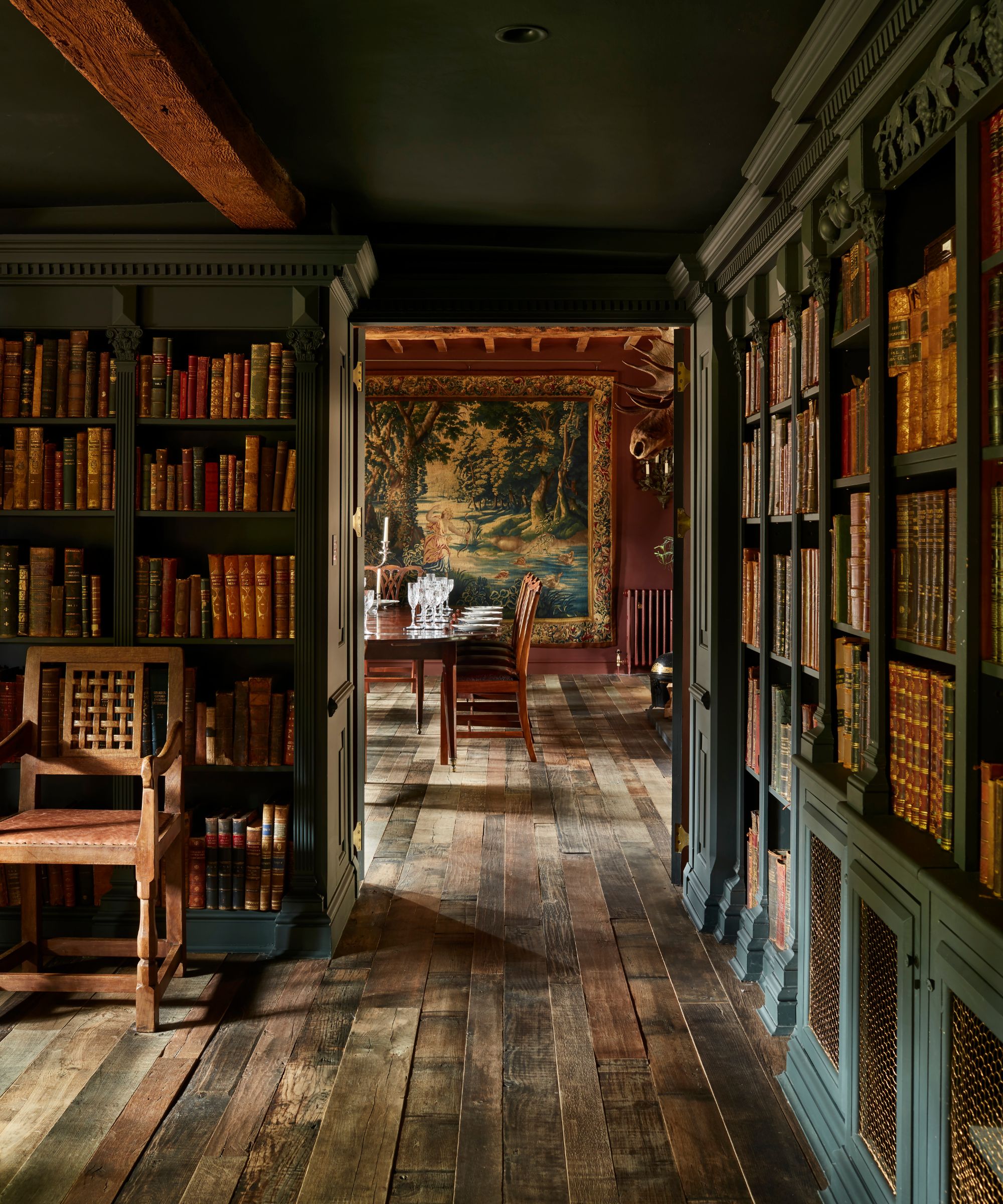 A dark-toned library with floor-to-ceiling green bookshelves filled with leather-bound books, leading into a formal dining room with a large wall tapestry and a wooden table