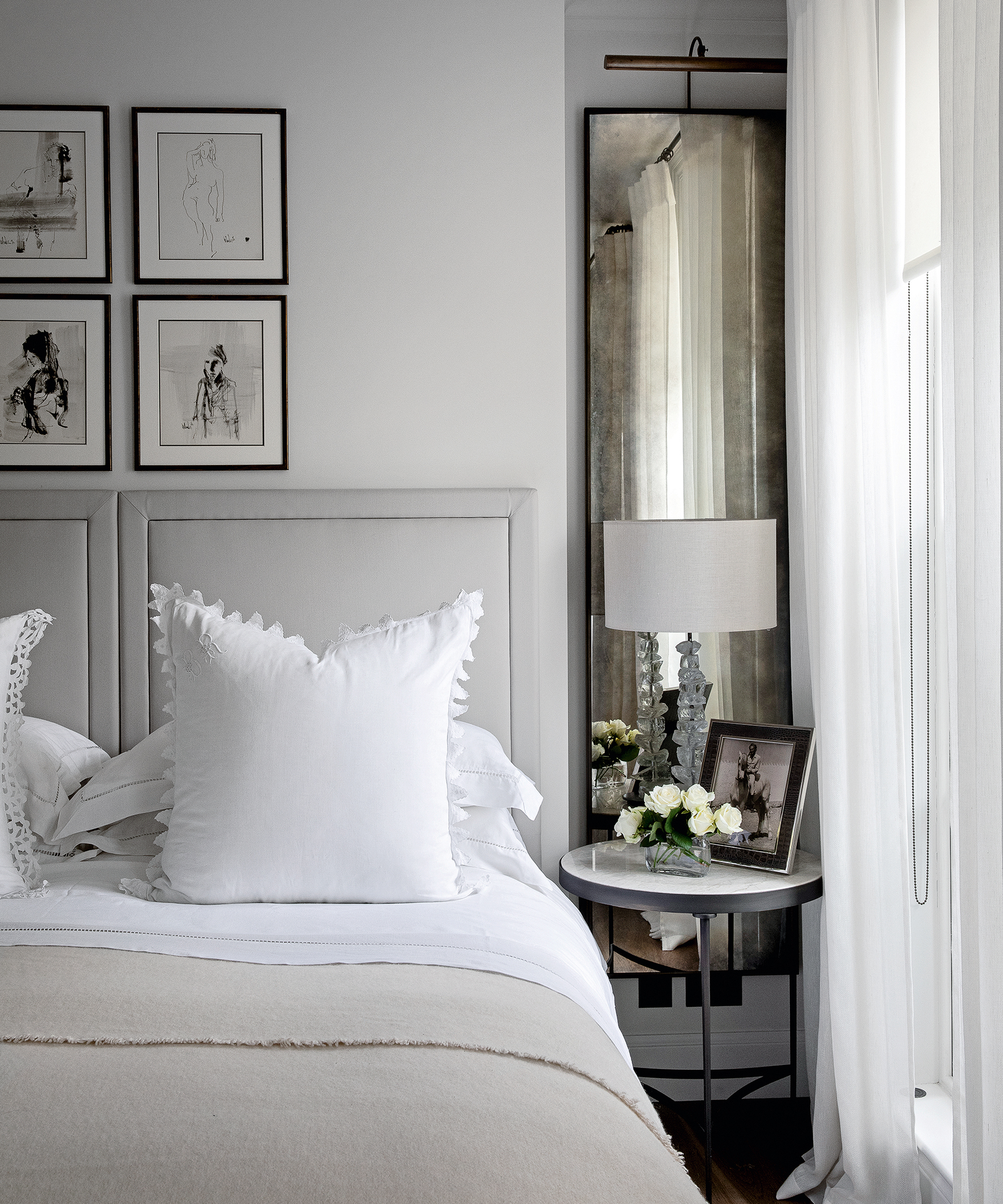 A bedroom color idea with white walls, dove grey headboard, white curtains, cream throw and tarnished black-framed mirror