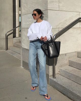 Influencer Sasha Mei poses outside a city building wearing aviator sunglasses, a long-sleeve white sweatshirt, baggy straight-leg jeans, a black top-handle bag, and blue-and-black flip-flop kitten heels.