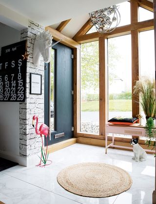 a large porch area with wooden beamed windows, a large black door, white tiled floor and fun accessories including a pink flamingo and pink side table with plants and a cushion, and a round jute rug