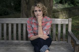Charlotte Ritchie as Bonnie, wearing a red and blue plaid flannel shirt and indigo jeans with big turn-ups, sitting on a bench resting her chin on her hand