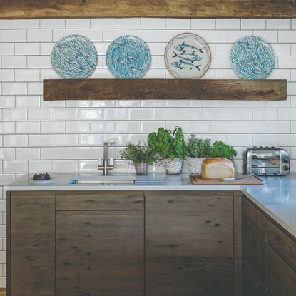 A white-tiled kitchen with potted herbs on the worktop and fish-motif plates displayed on a rustic shelf