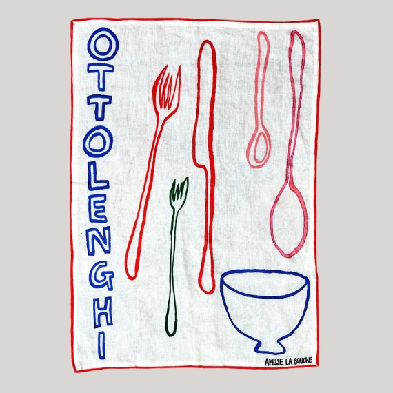 A white, blue, red, and green tea towel with handpainted tableware on it and the writing "OTTOLENGHI" sits against a pale grey background.