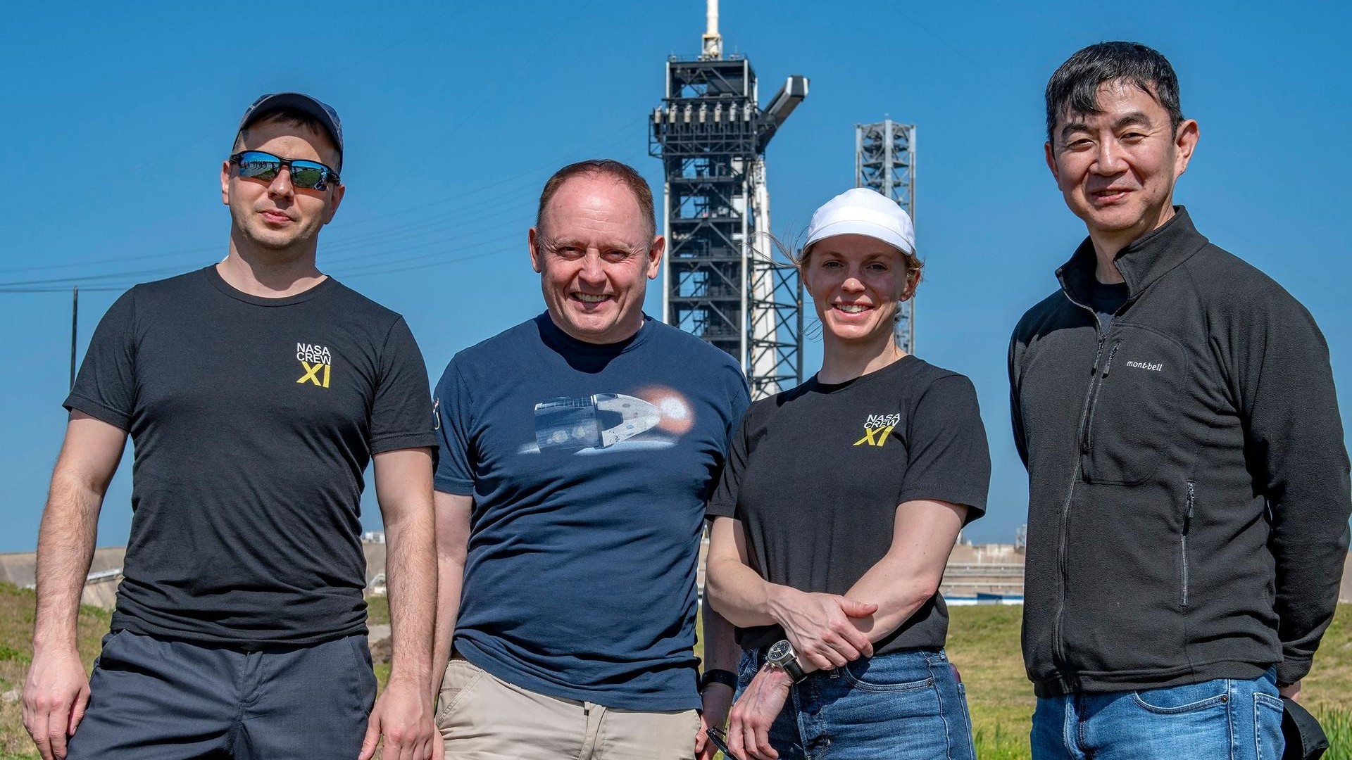 four people in t-shirts pose under a blue, sunny sky in front of a rocket on a launch pad