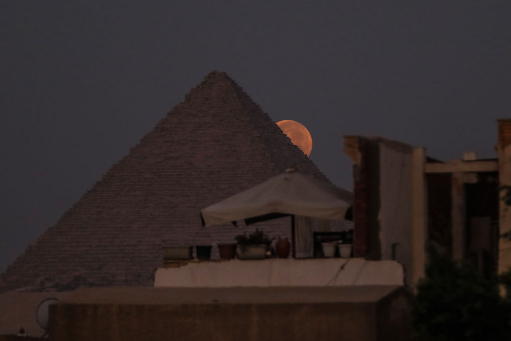 the rosy red/orange hued full moon peeks out from the side of a large pyramid.