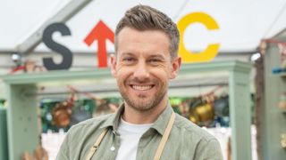 Chris Ramsey wearing a green shirt and bake off apron, smiling at the camera