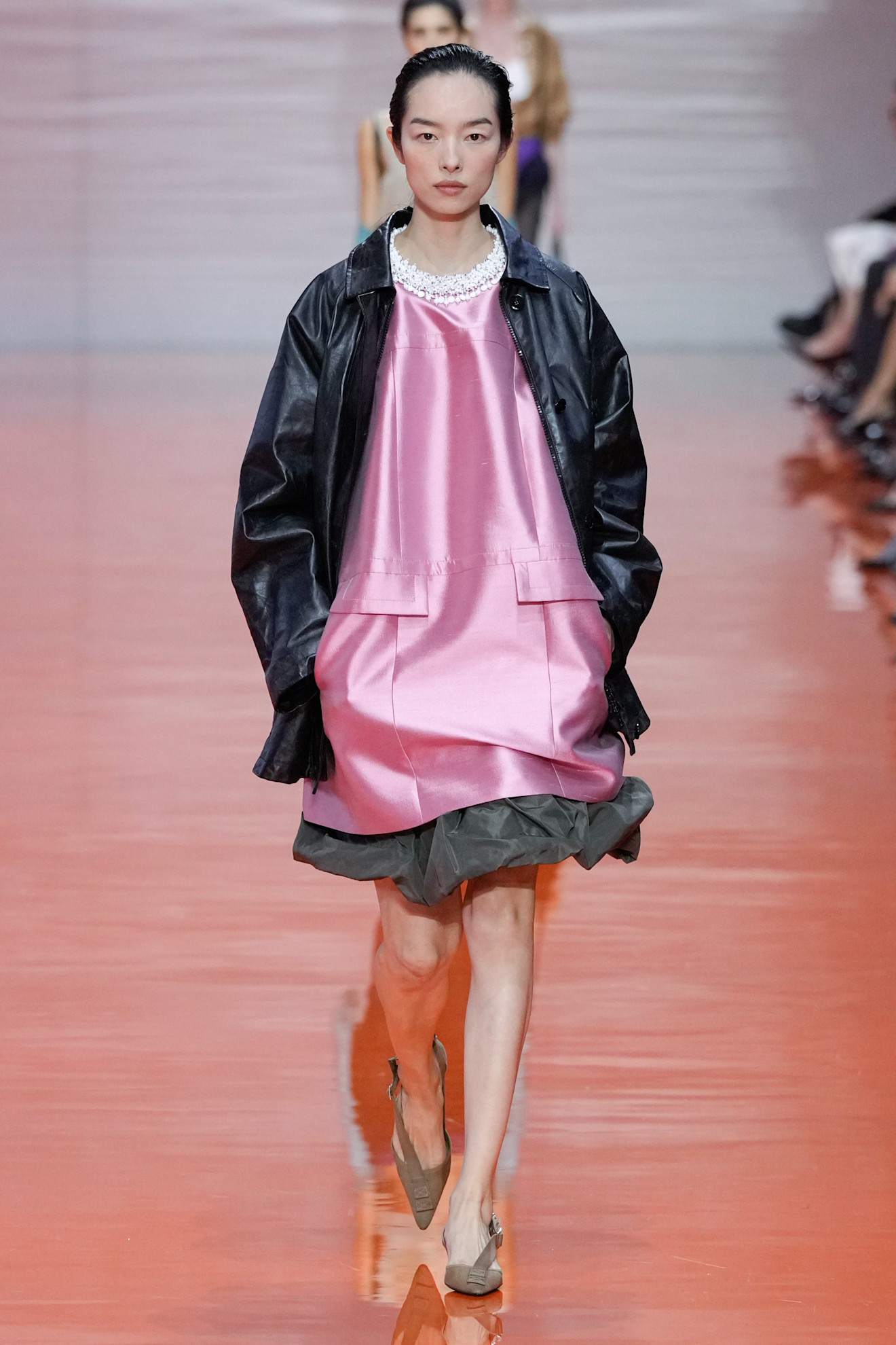 A model walking in the Prada SS26 show wearing a satin shift dress with a jewel collar and a leather jacket.