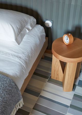Side view of a bed and a round wooden side table with different coloured grey floor tiles.
