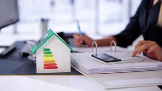 model of house with energy ratings on side, woman in background using calculator resting on folder of papers