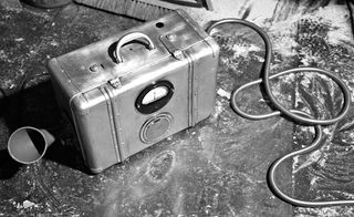 Black and white image, suitcase with electrical dial, side spout, metal cord, speckled concrete floor, sweeping brush head in shot