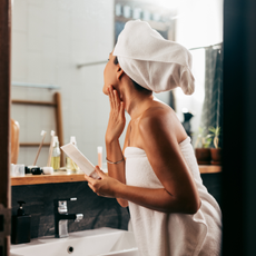 Woman Wrapped In A Towel Looking Herself In The Mirror While Applying Some Creme On Her Face After Taking A Bath