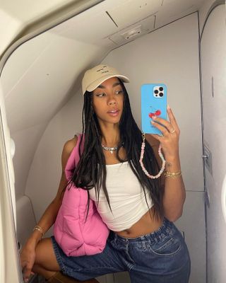 Dutch fashion influencer Amaka Hamelijnck taking a selfie on an airplane wearing a white tank, a baseball hat, denim shorts, and a pink tote bag.