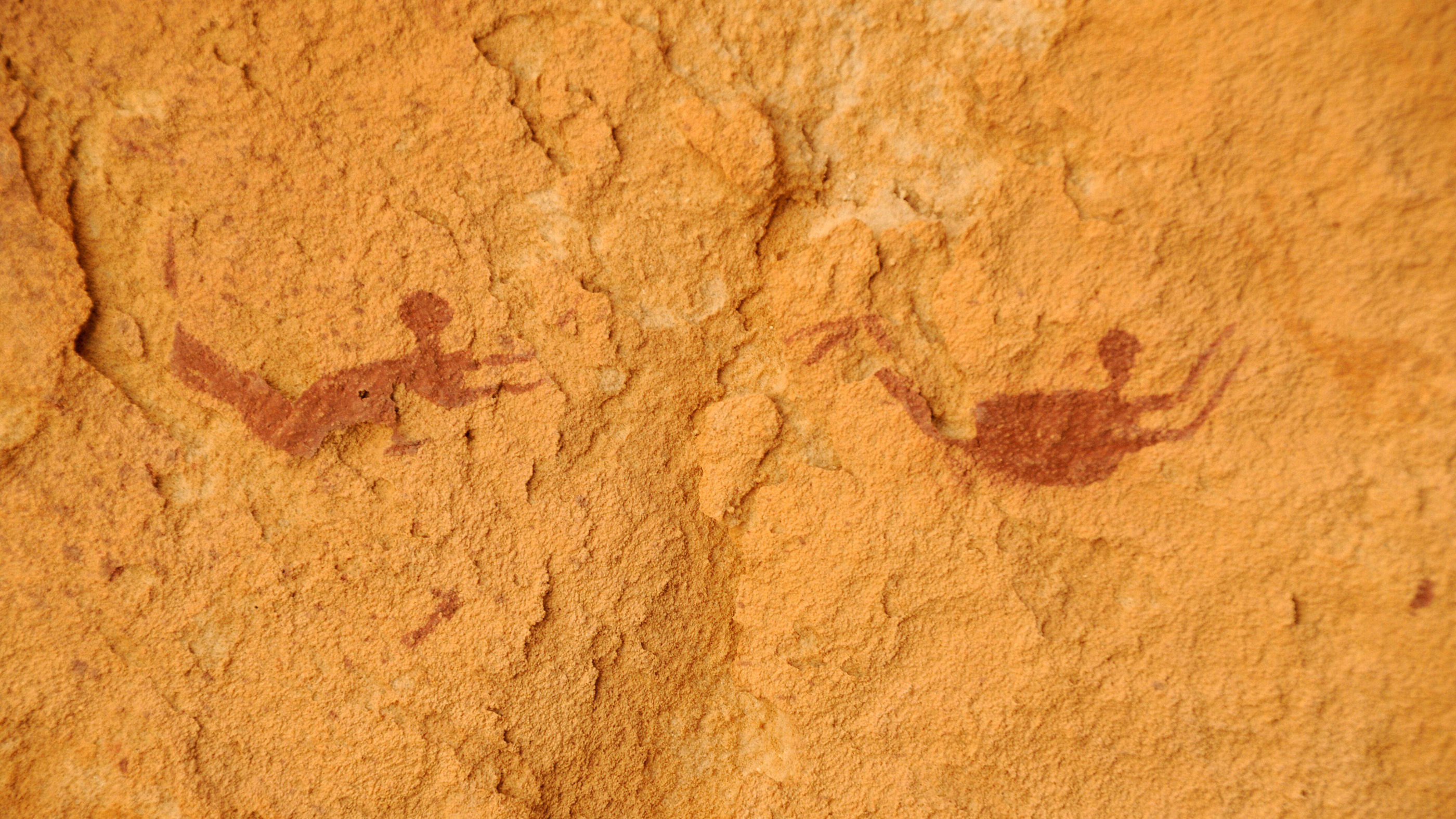 A close-up showing two red paintings of human swimmers against an orange-y background in the cave of swimmers.