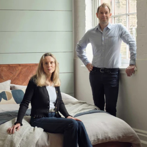 Image of a woman sitting on a bed and a man standing beside the bed by a window.