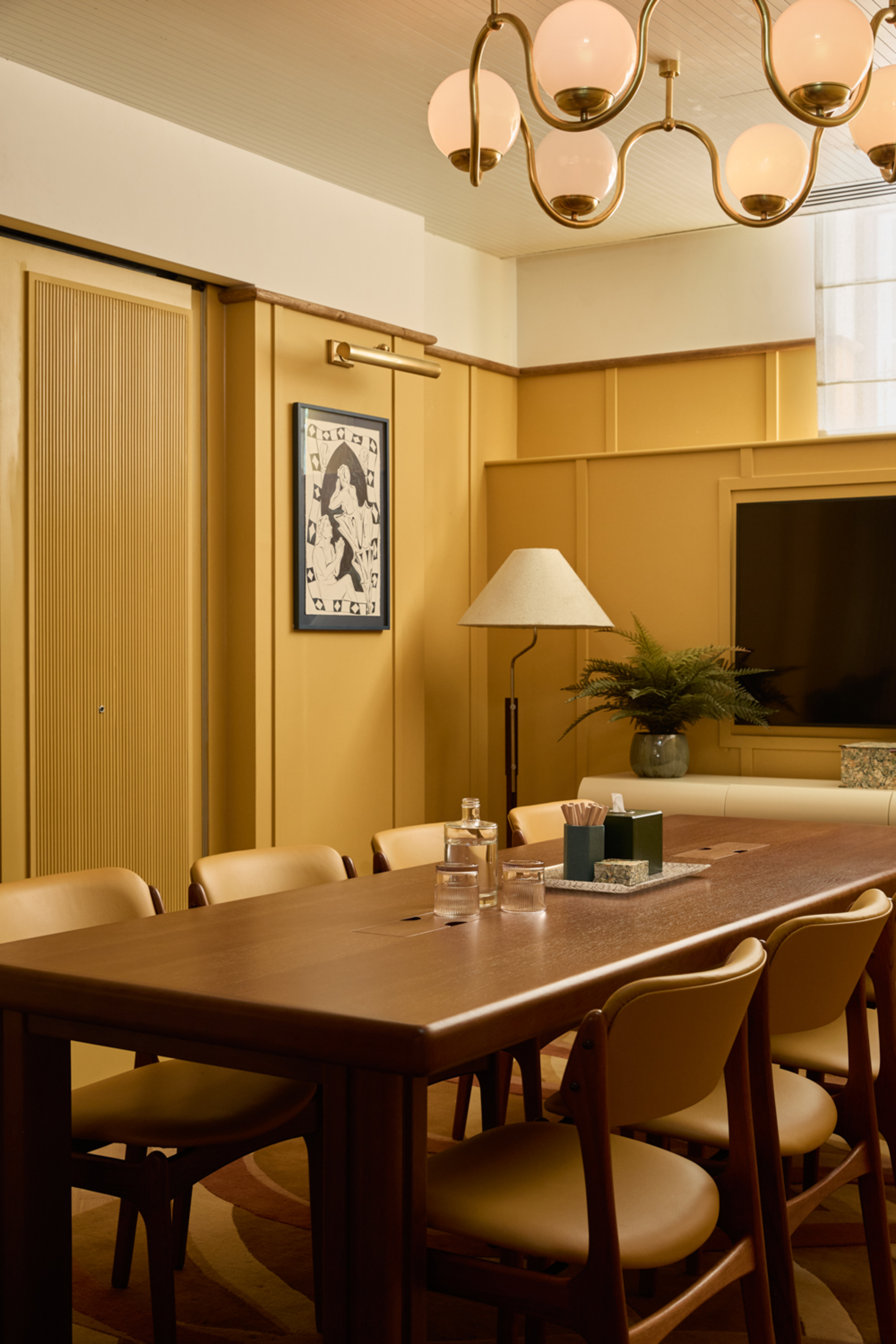 dining room with yellow wall paneling, floor lamp in corner and wiggly pendant light above timber table with leather dining chairs, a TV at the head of the table with a console table with a potted plant