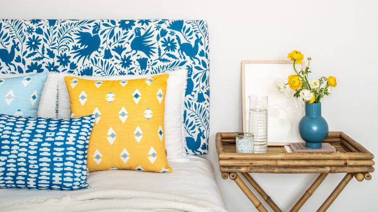 Blue patterned upholstered headboard with patterned yellow and blue tone scatter pillows, and bamboo bedside with yellow flower posy and water carafe.