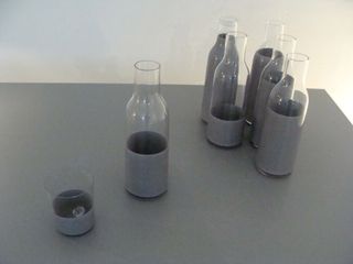 Five two-toned (bottom: grey and upper: glass) bottles and a two-toned (bottom: grey and upper: glass) tumbler photographed on a grey table against a white wall