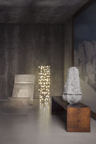 In a dimly lit gallery with grey, stone-like walls, linear, futuristic furniture items including a deck chair, a standing lamp, a bench, and a sculpture stand next to each other.