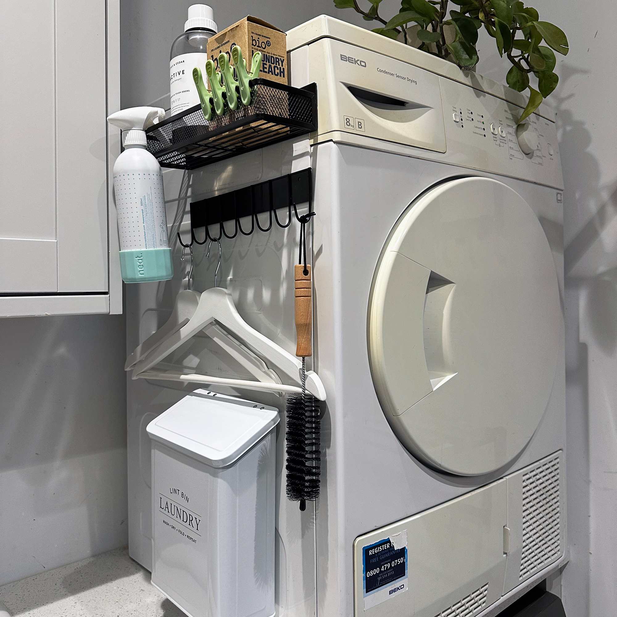 White tumble dryer stacked above a washing machine in a white utility room, with black magnetic shelves from Amazon lined up on the side