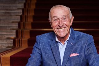 Len Goodman wearing a blue check blazer and a pink pocket square, sitting on a red carpeted stairway