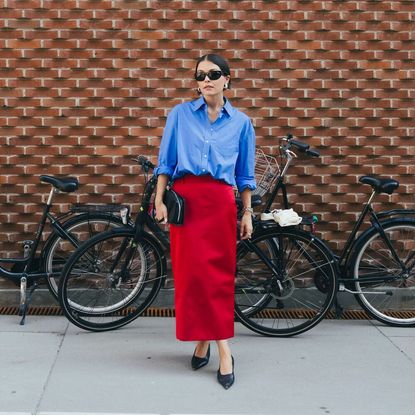 Woman wears red skirt, blue button down, and black heels while posing in front of many bikes. 