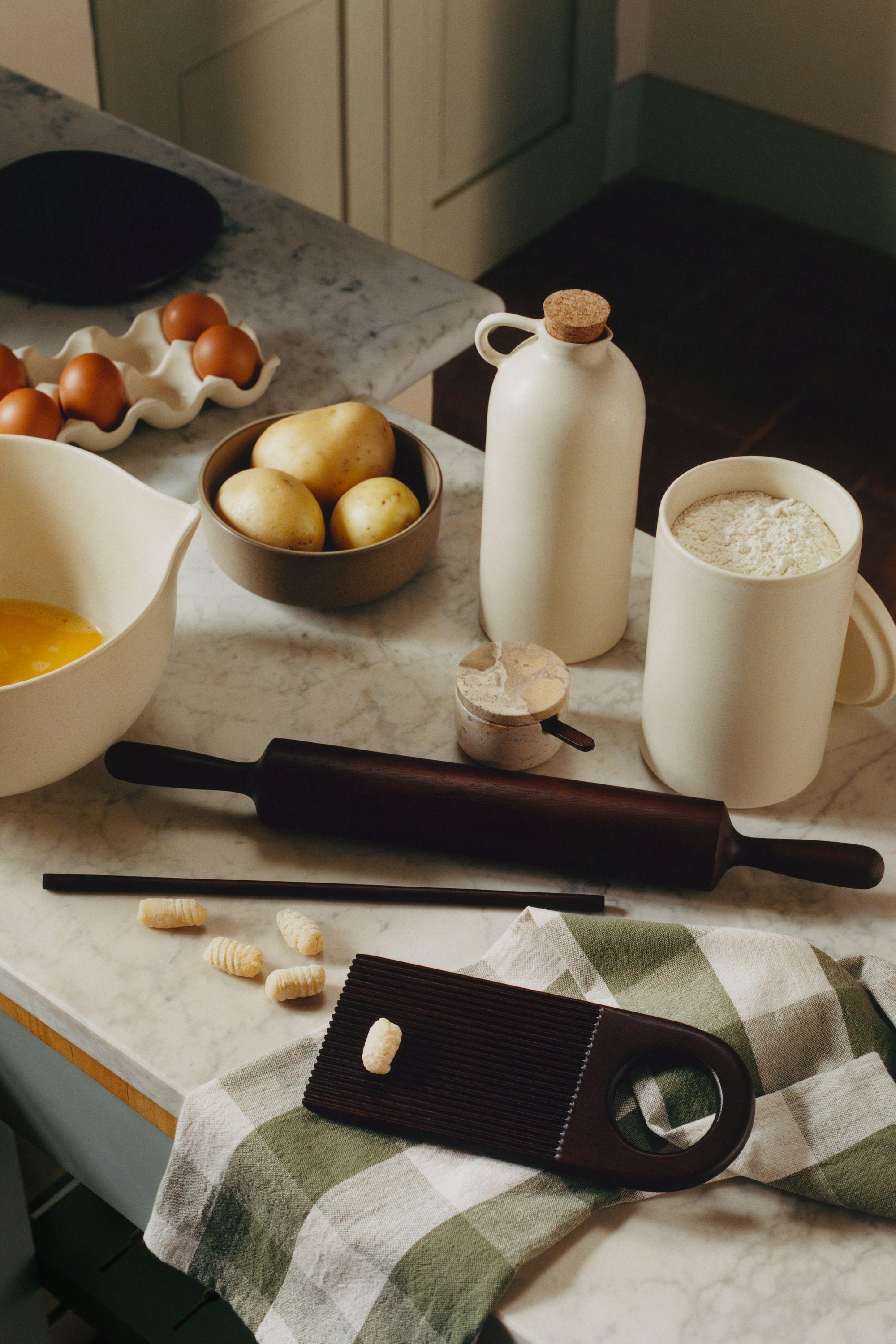 Close-up image from above of a marble kitchen countertop with different cooking utensils on it. There is a dark wood rolling pin and gnocchi making set, A ceramic jar of flour, a bowl of potatoes, and a ceramic egg holder.
