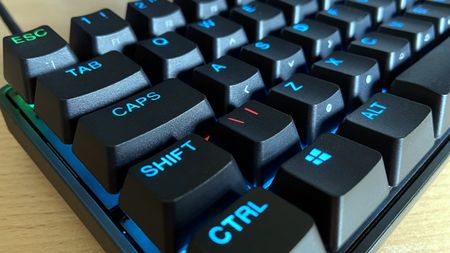 Close-up of a black mechanical keyboard with illuminated blue-lettered keys like "CTRL" and "SHIFT" on a wooden surface, creating a modern tech vibe.