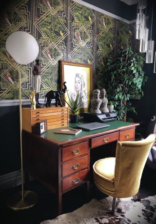 a traditional but eccentric home office with a yellow chair, a vintage wood desk, jazzy green wallpaper, and art balanced on the desk