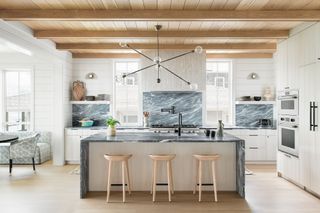 beadboard kitchen cabinets in pale colors with timber ceiling and floors and a large marble island in the middle of the kitchen with bar stools.