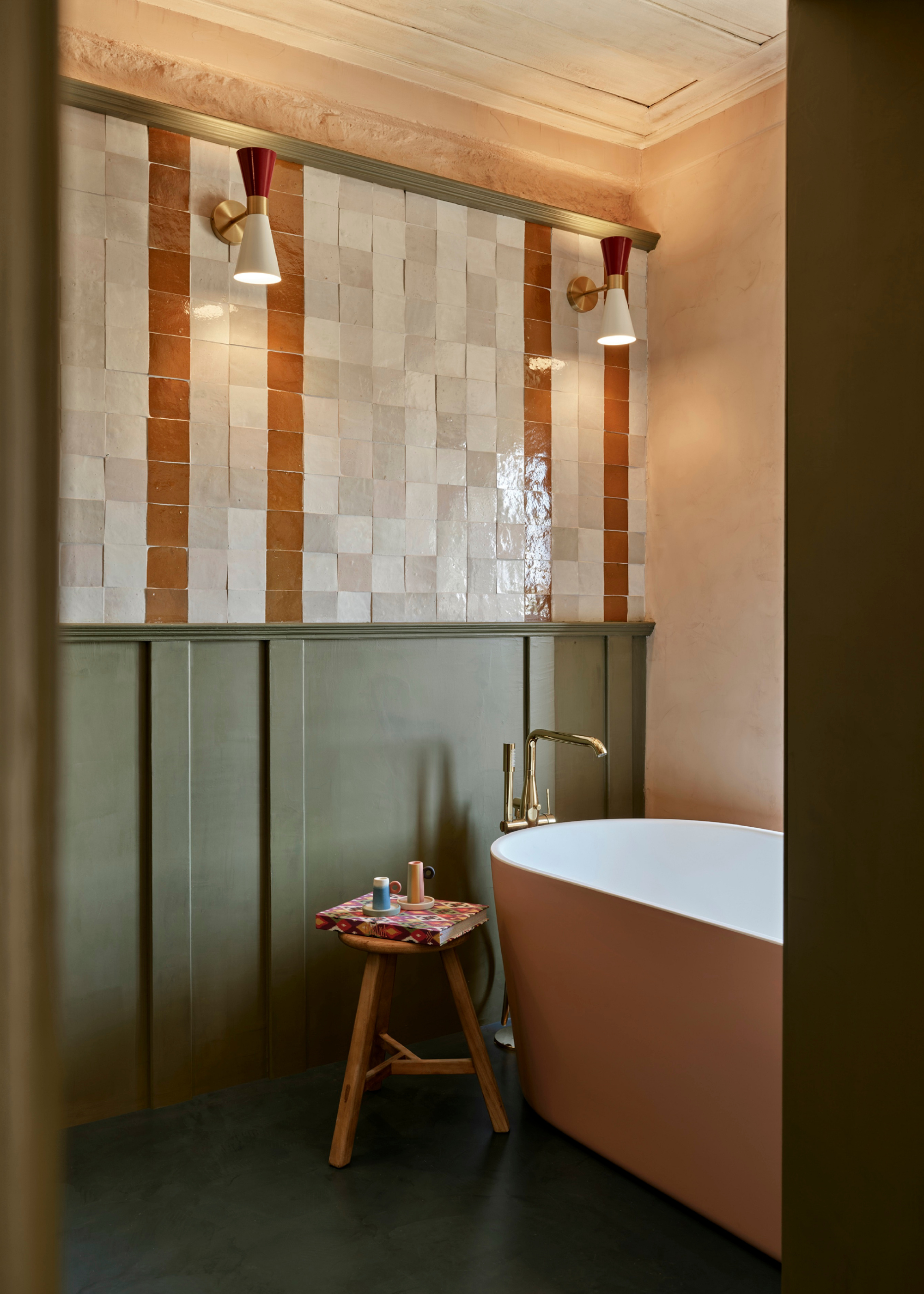 a bathroom as seen through the door frame. Free standing bath with a wooden stool besides it. the walls have zellige tiles on the upper half, and wooden panelling beneath. the floor tiles are a dark green tone, matching the panelled walls.