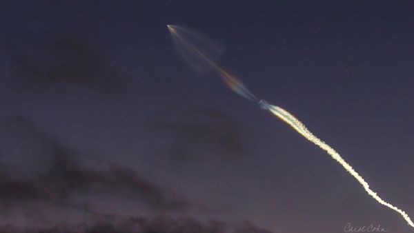 a trail of smoke wisps far away in the sky, tracing upward to a rocket. it is nighttime 