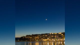 two bright orbs in a deep blue sky above the lights of a small town on a hillside, the lights from which are reflected in a lake