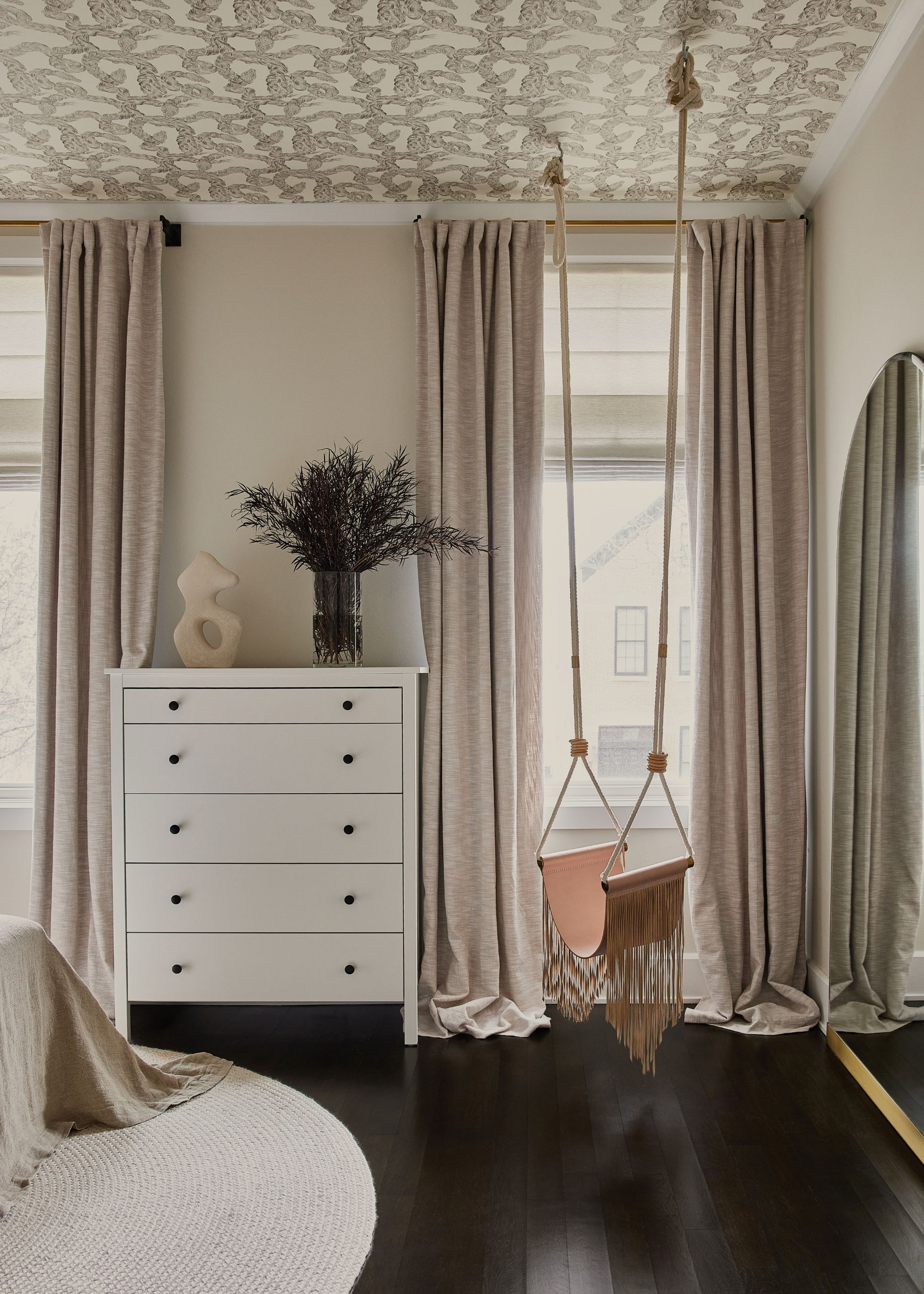 A corner in a bedroom with dark flooring, a white chest of drawers and a swing with a fringe design