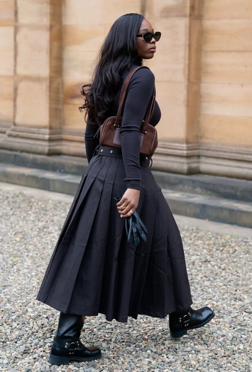 @rabia.cissokho wears a black long-sleeved top, pleated skirt, biker boots and brown shoulder bag
