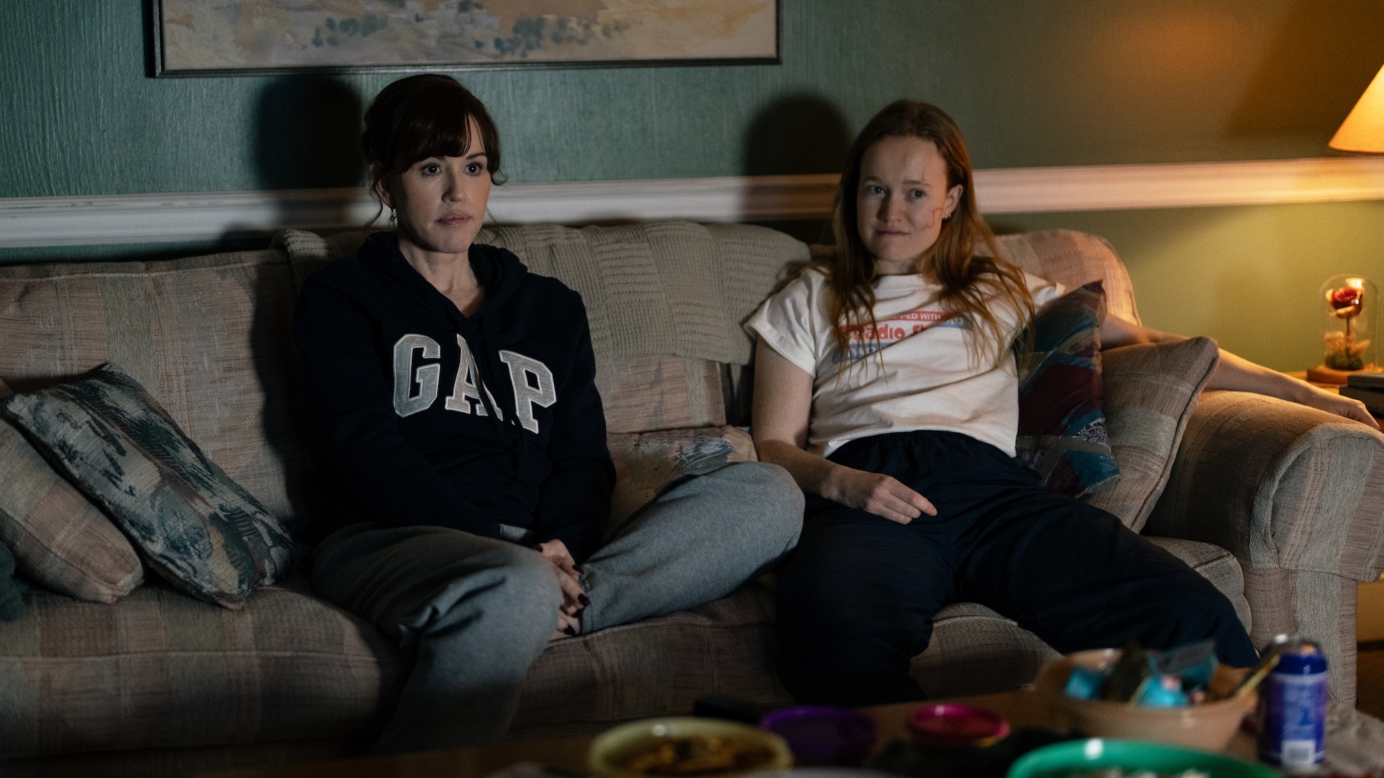 Vicky Palmer in Gap sweatshirt and sweatpants sitting on sofa with Van in front of a snack-filled coffee table while watching TV in Yellowjackets Season 4