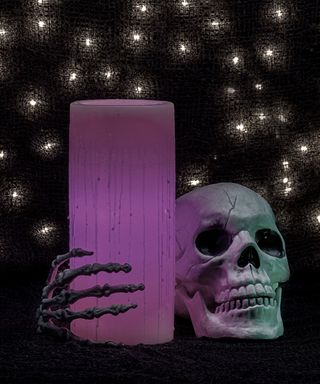 A purple candle being held by a faux skeleton hand beside a faux skull on a black background
