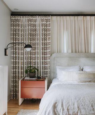 Mono patterned and plain full length drapes behind upholstered gray bed with relaxed bedlinen, and black wall light.