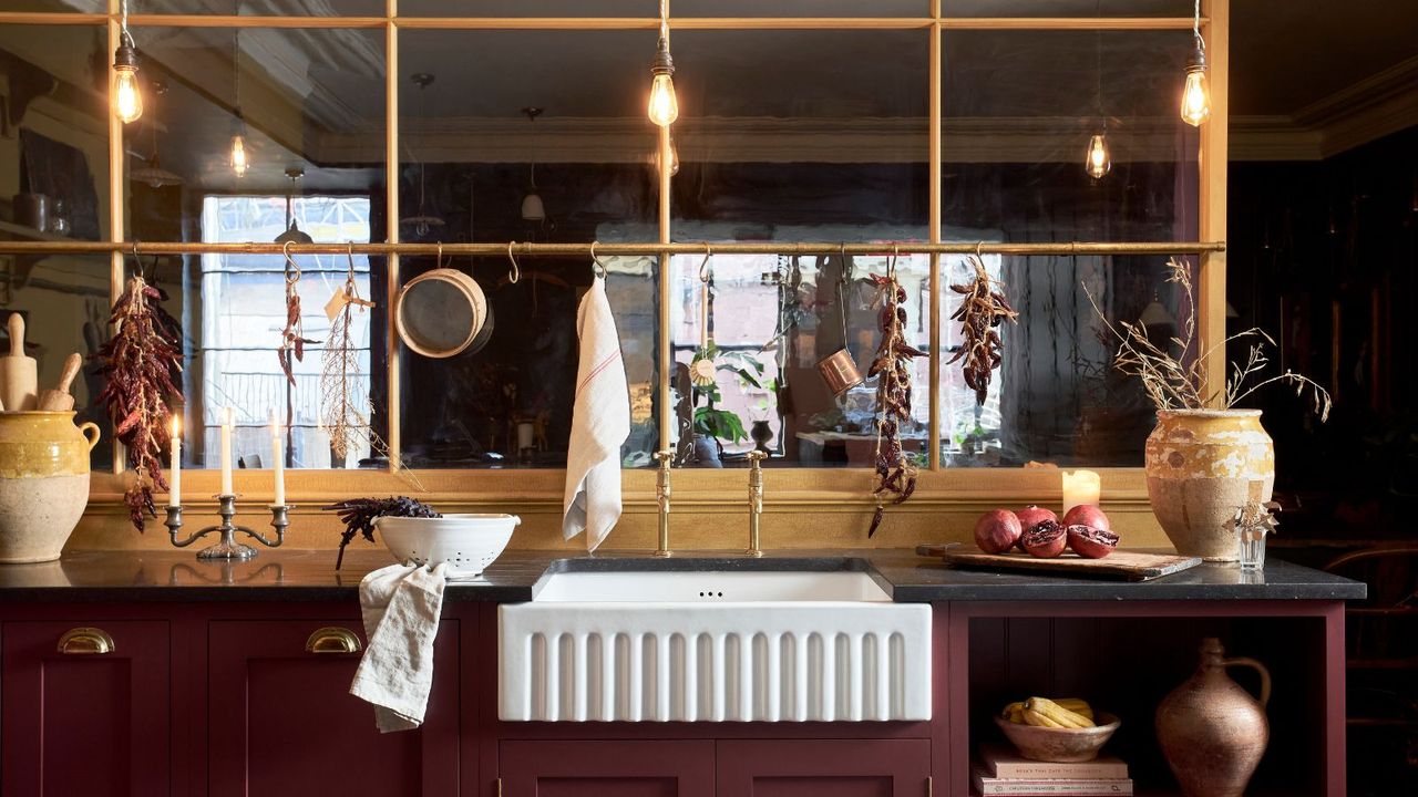 A burgundy, wood and brass kitchen with glass paneling, dried herbs and flowers, and black marble countertops with contrasting white sink