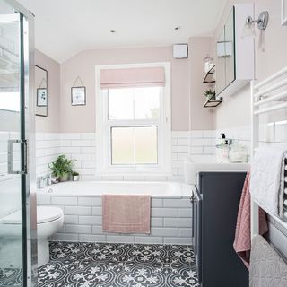 a bathroom with top half of wall in blush pink paint and bottom half with white tiles, a white tiled inbuilt bath, a black and white patterned floor and black undersink cabinet