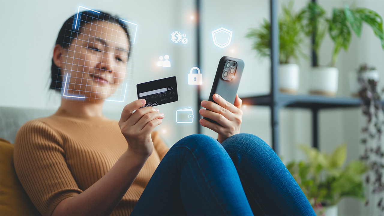 Woman sitting on sofa in living room at home holding credit card and scanning face id on a smart phone 