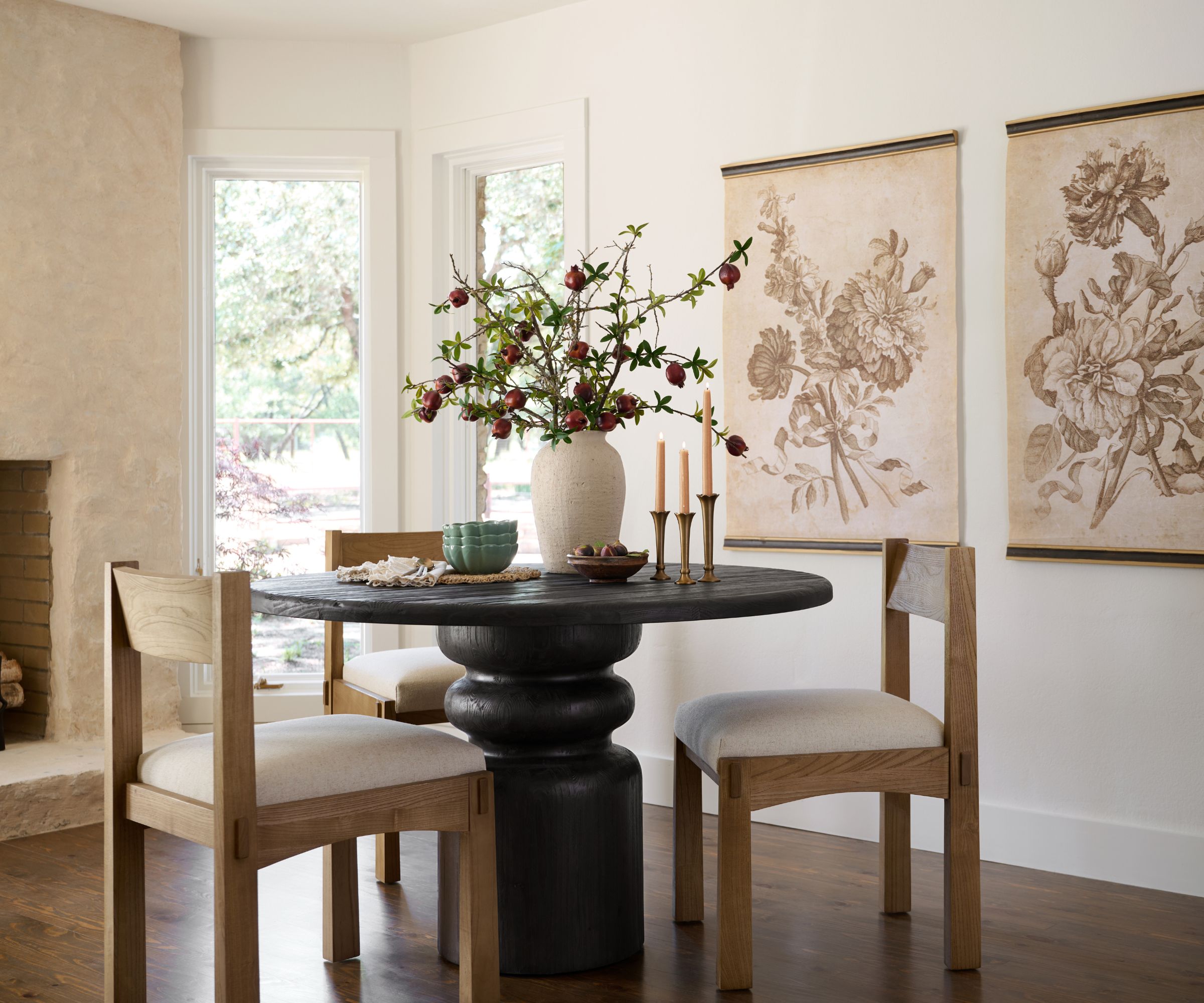 Dining room with curved dark wood table, light wood dining chairs, vintage prints on walls of flowers, large white vase with flowers and gold candlesticks