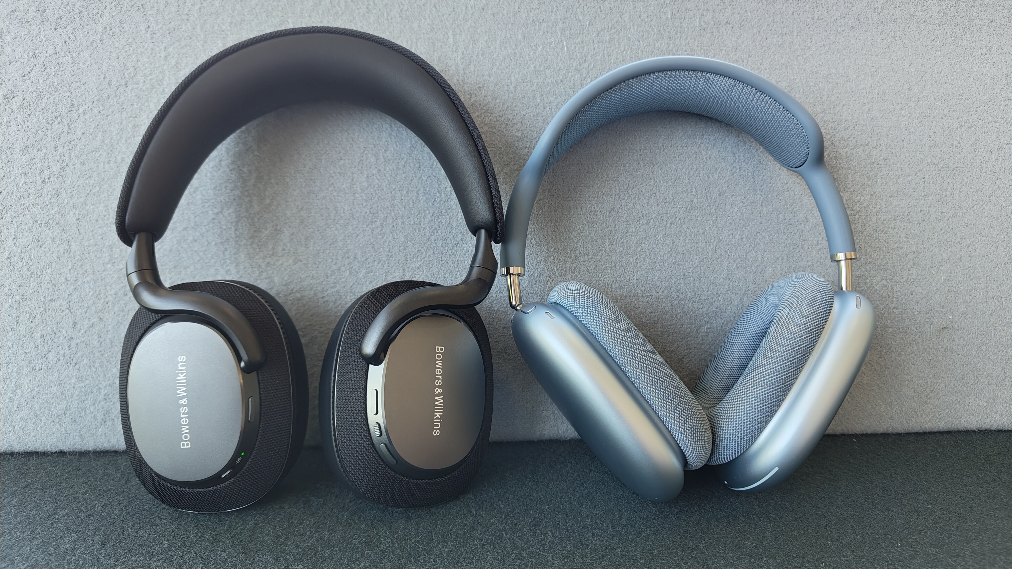 A black pair of B&W Px& S3 headphones (left) propped up against a wall next to a pair of blue Apple AirPods Max (right).