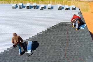 Two workers on a roof adding a new layer of roofing tiles.