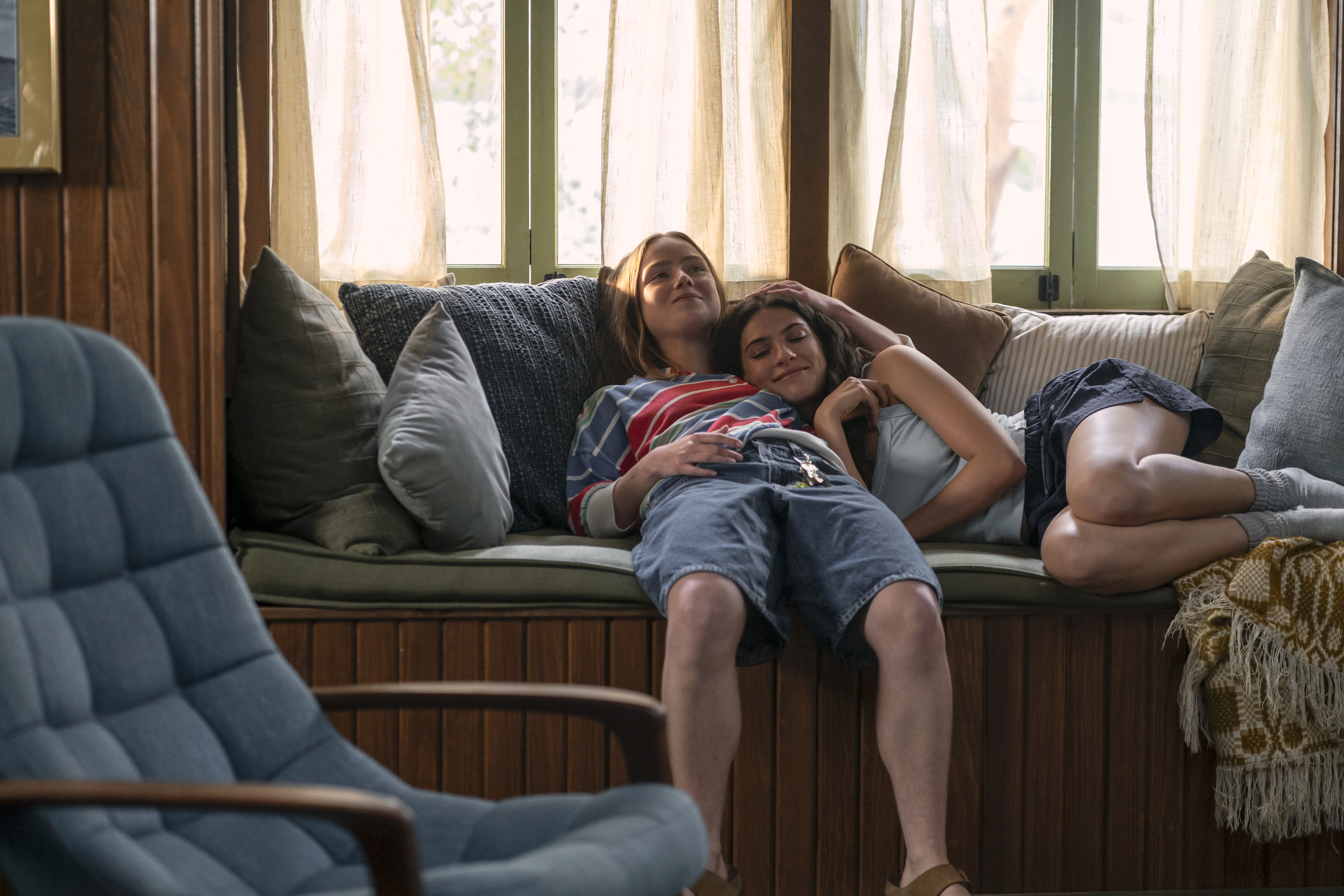 Two teen girls (Ame&amp;#769;lie Hoeferle as Ramona and Ella Rubin as Annie Jacobson) lie together on a bench in a cottage living room, in the Prime Video series 'Sterling Point.'