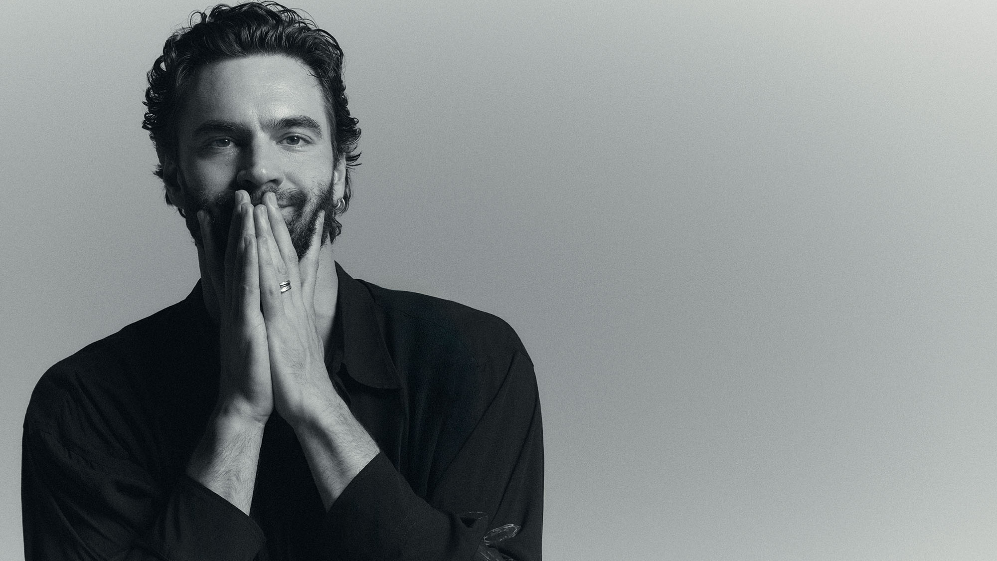 Portrait of Tom Bateman shot by Graham Dunn. Wearing a navy button-down shirt, he poses with his hands in front of his mouth. 