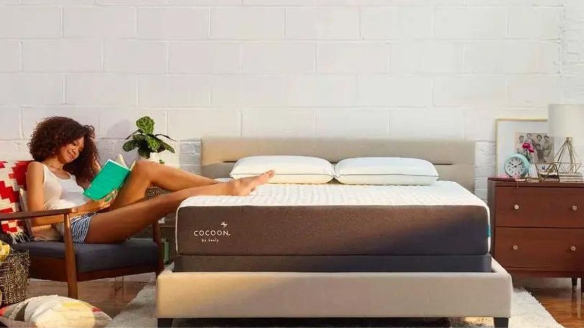 A woman wearing a white singlet an d denim shorts sits in a chair with her legs restiong on top of the Cocoon by Sealy Chill mattress