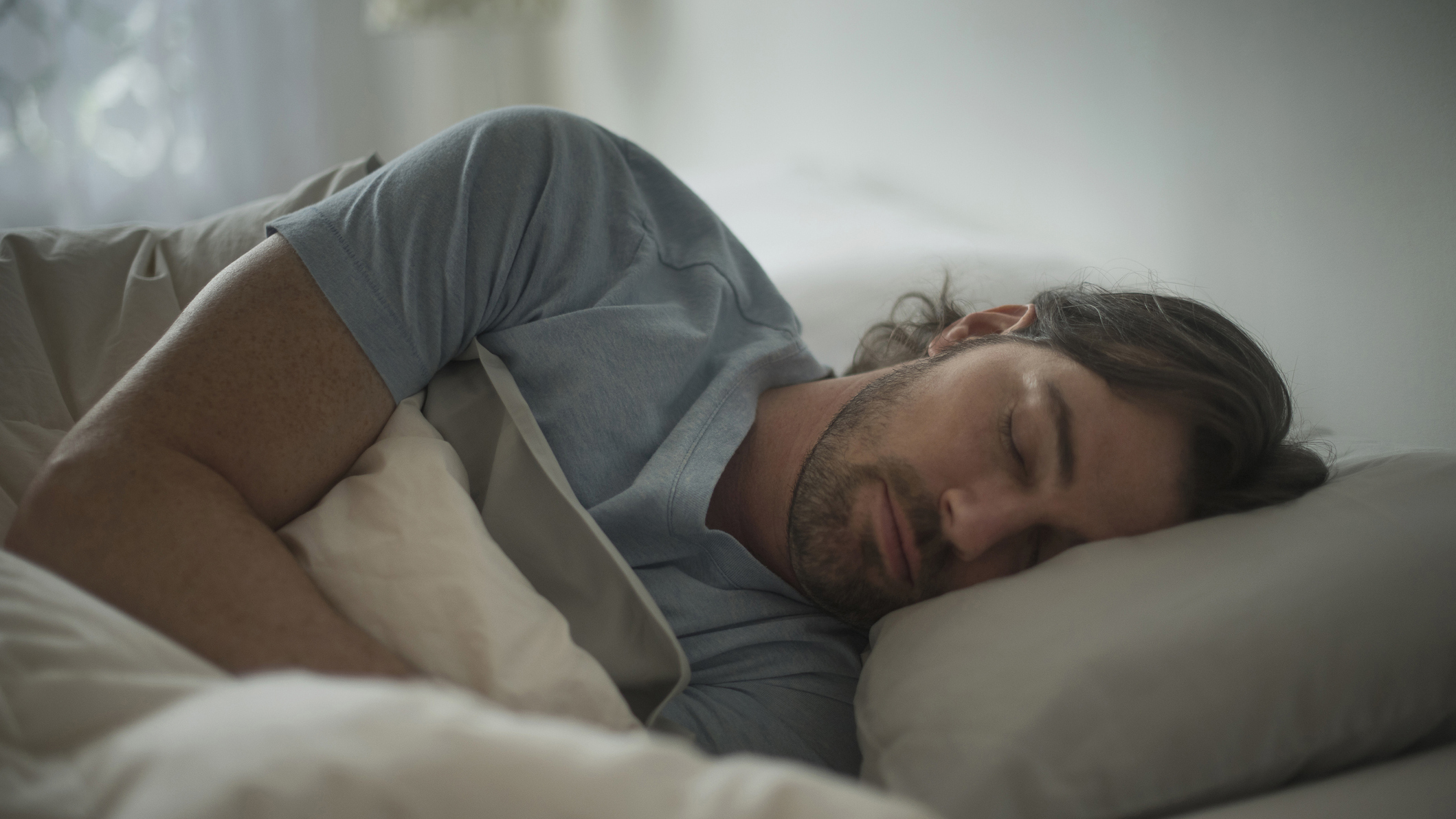 A man with long dark hair sleeps on his side in bed to reduce his chances of snoring