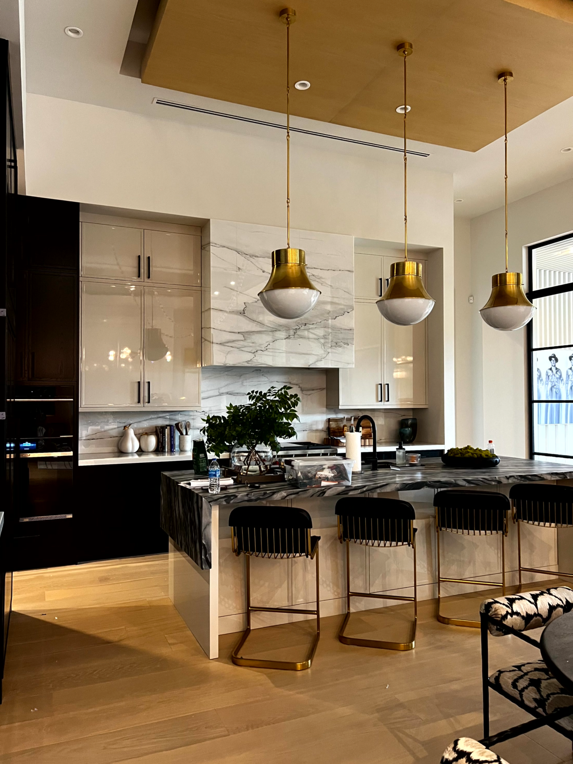 A luxury kitchen with a large island, three gold pendant lights, glossy cabinets, and a marble hood