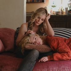A blonde woman (Elle Fanning) lies on a pink couch with black-and-white striped pillows, resting her head on a pillow in her mother's (Michelle Pfeiffer) lap. The pair sit in a living room with a kitchen behind them, through a circular doorframe, in Apple TV+'s 'Margo's Got Money Troubles.'